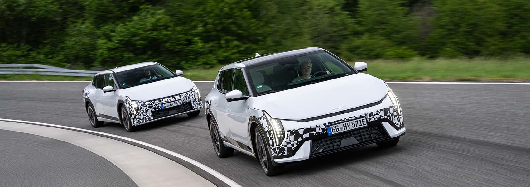 Dva bílé maskované modely elektromobilu Kia EV4 projíždějí zatáčkou na testovací dráze.