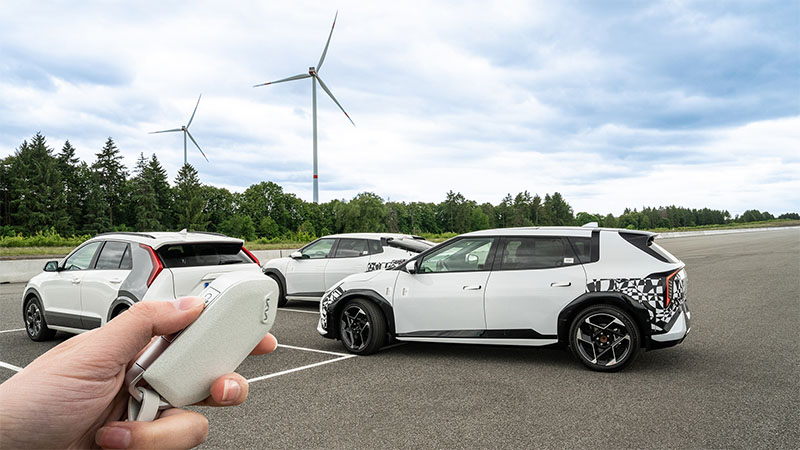 Modely elektromobilu Kia EV4 a Kia Niro EV na zkušební dráze. V pozadí jsou vidět větrné turbíny