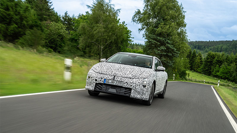 Kia EV4 s maskovacím vzorem jede po silnici v přírodním prostředí