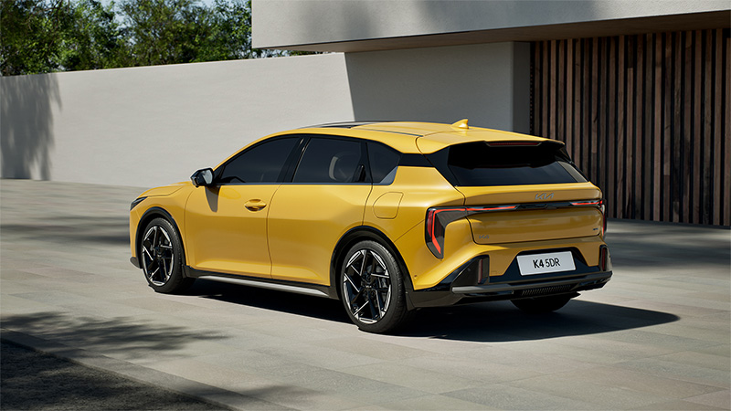 Žlutý model Kia K4 projíždějící kolem domu - boční pohled zezadu