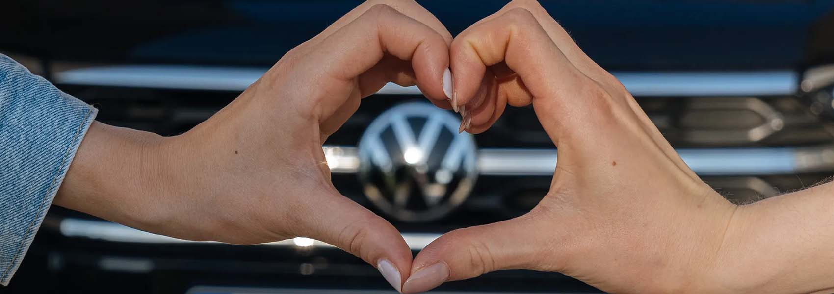 Dvě ruce tvořící tvar srdce před maskou auta s logem Volkswagen.